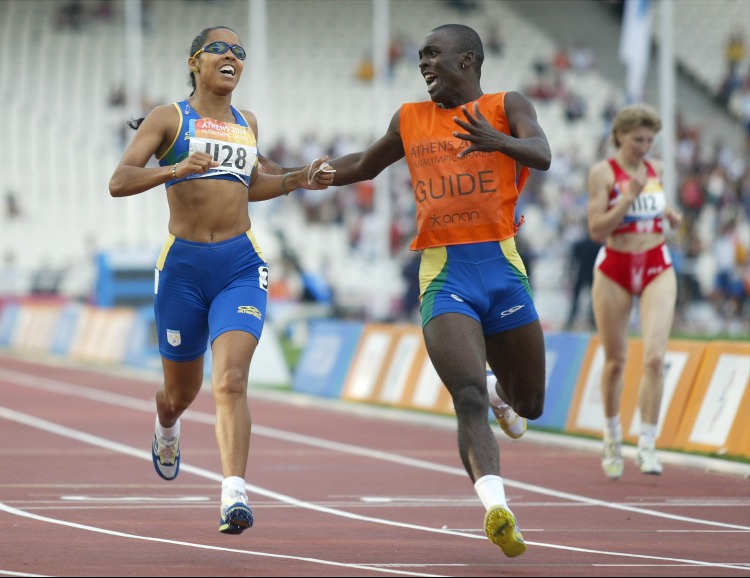 A esquerda, Adria veste uniforme azul do Brasil e usa óculos de sol preto. Adria está acompanhada de seu atleta-guia da época, que veste camiseta laranja e short azul com detalhes em amarelo e verde. 