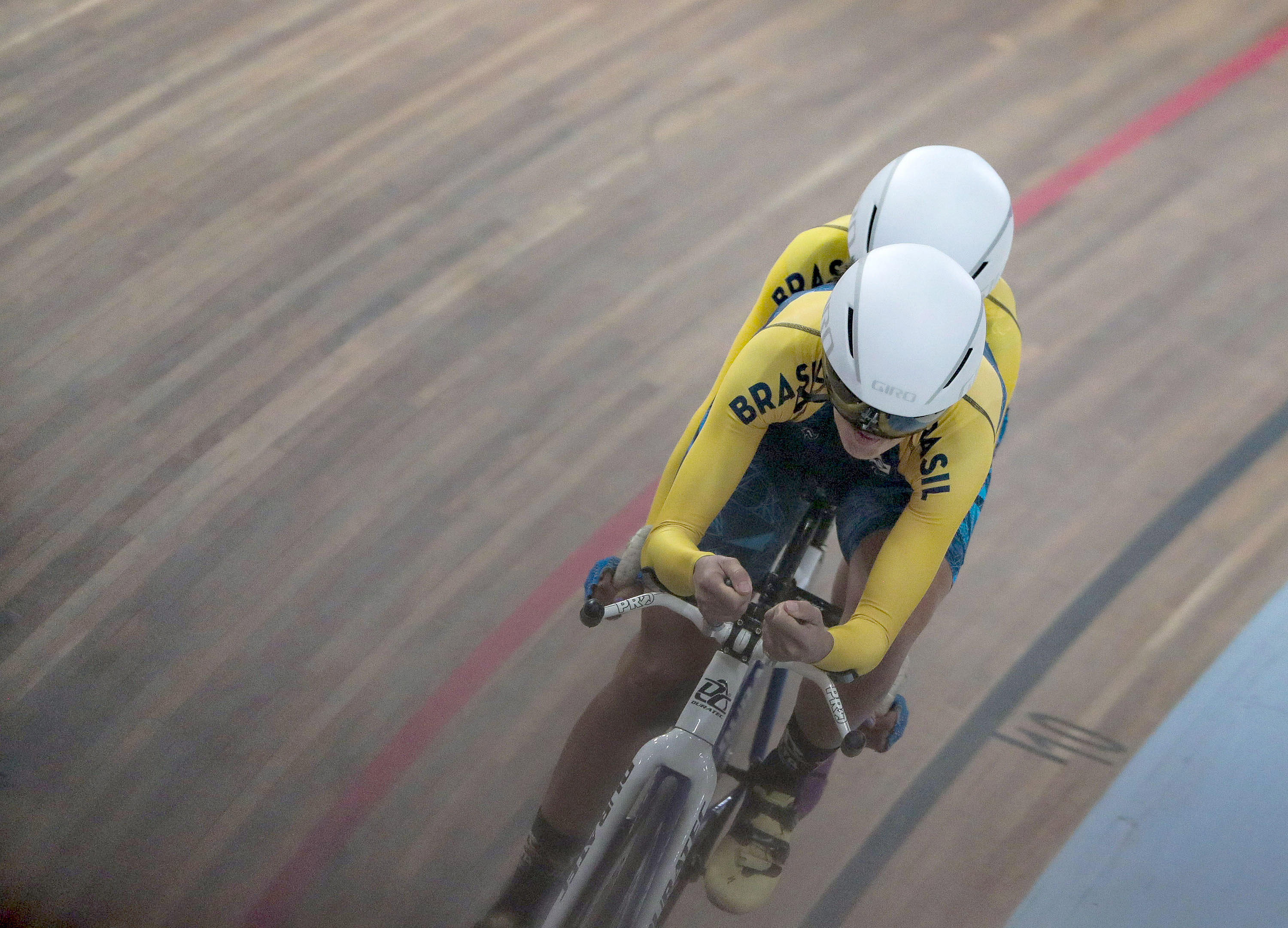  Na foto, vê-se dois ciclistas em uma bicicleta tandem branca durante uma prova em um velôdromo com pista de taco marrom e uma faixa vermelha; um ciclista está atrás do outro e ambos vestem o uniforme do Brasil, camiseta amarela e capacete branco.