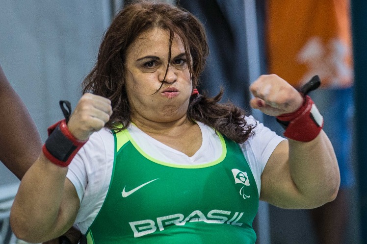 Márcia está com as duas mãos fechadas e levantadas a altura do rosto. A atleta utiliza macacão do Brasil verde e por baixa uma camisa de manga curta branca. Nos punhos, ela usa uma faixa vermelha em cada.