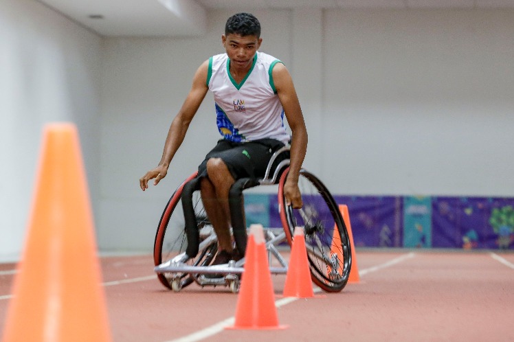Foto horizontal de um jovem negro em uma cadeira de rodas correndo entre cones laranjas em uma pista interna de atletismo.