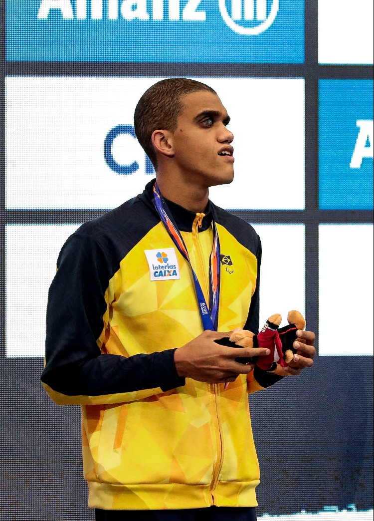 Na foto, o nadador Wendell Belarmino, de pele morena e cabelos raspados, segura bichinho de pelúcia durante premiação de natação. Ele veste jaqueta amarela e azul marinha da delegação brasileira e tem medalha pendurada no pescoço.
