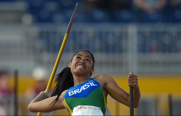 Foto de Raissa em uma pista de atletismo. Ela segura um dardo na mão direita e apoia a outra mão segura uma haste de apoio. Raissa está com o tronco levemente inclinado para trás. Usa uniforme do Brasil nas cores verde, amarelo e azul.