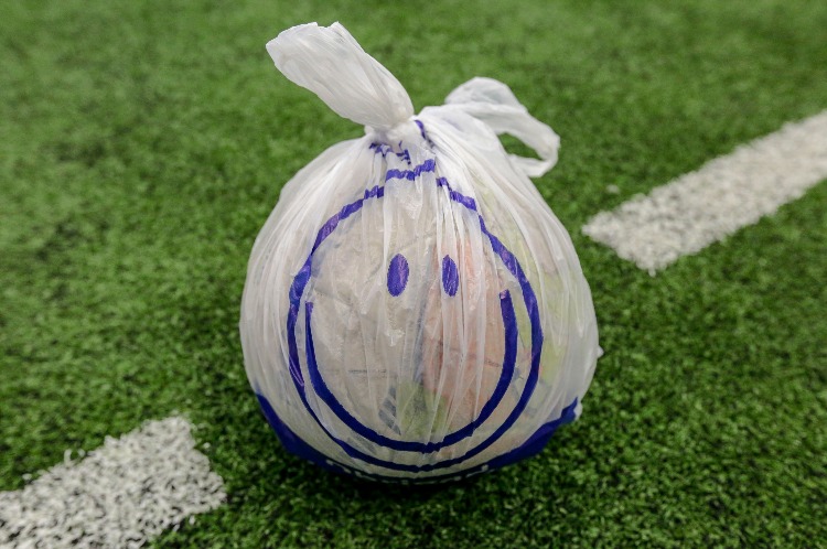 Em destaque na foto, está uma bola dentro de uma sacola branca com um nó em cima e com o desenho azul de um rosto com dois olhos e um sorriso. A bola está sobre um gramado sintético verde com uma faixa branca.
