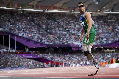 Alan Fonteles, homem branco, usa óculos de sol preto, uniforme do Brasil com bermuda verde e blusa com detalhes azul e amarelo. Alan é biamputado de pernas e utiliza duas próteses com lâmina de carbono. Ao fundo, o estádio dos Jogos Paralímpicos de Londres 2012.