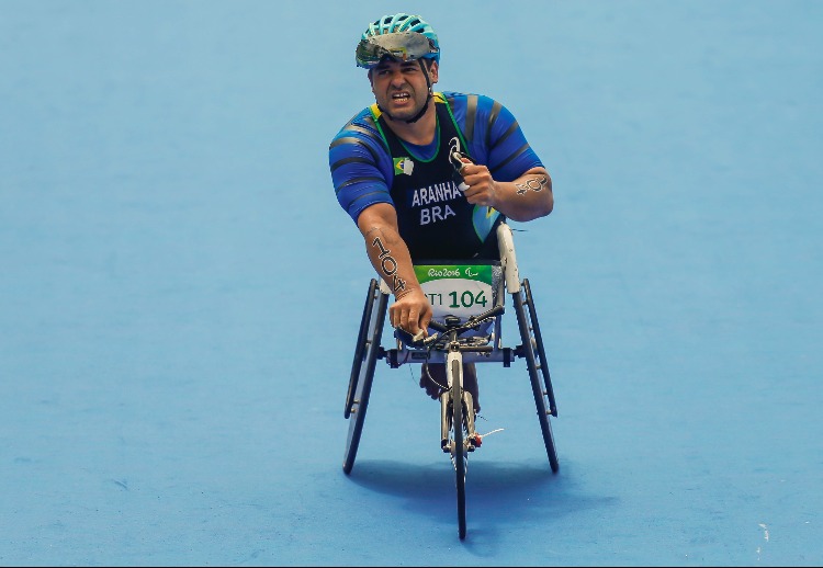 Fernando Aranha, homem, com capacete azul e preto, uniforme do Brasil azul com nome em letras brancas, na cadeira de corrida durante uma prova de triatlo