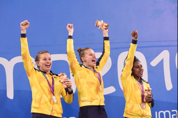 Da esquerda para direita: Beatriz Carneiro, Débora Carneiro e Ana Karolina Soares. Todas vestindo o casaco do uniforme do Brasil na cor amarela, com medalha pendurada no pescoço e braço levantado.