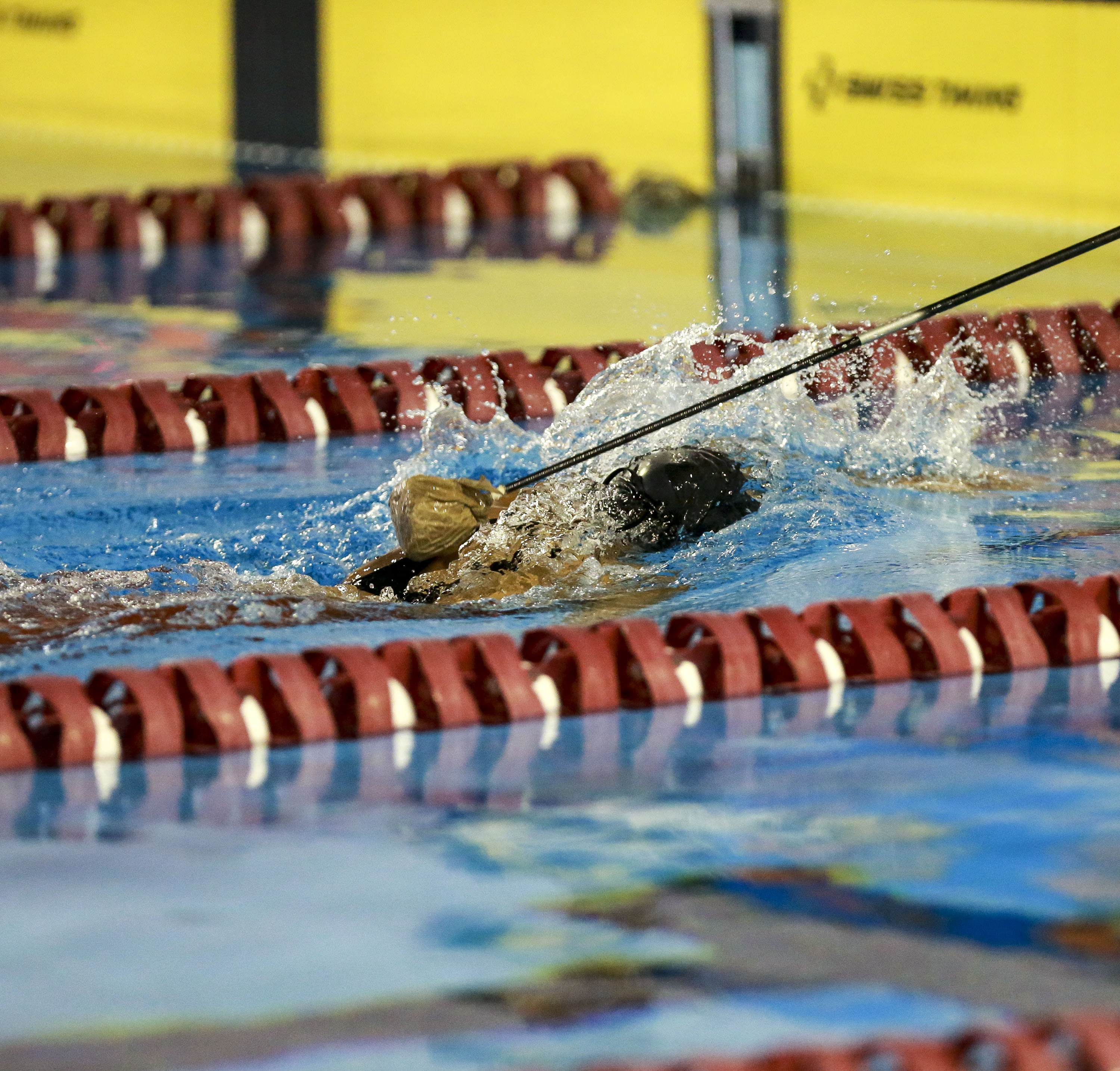 Na foto horizontal, um nadador com deficiência visual, usando uma touca preta, está proximo a borda da piscina com raias vermelhas e placas amarelas. O atleta recebe a sinalização do tapper, que é um bastão longo preto com esfera macia na ponta.