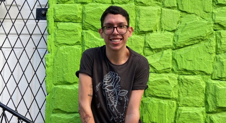 Ivan Baron em frente a um muro verde limão. Ele é um homem branco de cabelos castanhos escuros curtos, usa óculos com armação arredondada e sorri. Usa uma camiseta cinza  com estampa azul e vazada em formato de rosa.