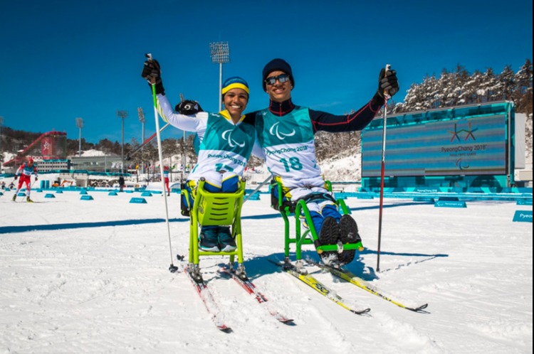 Atletas Aline Rocha e Cristian Ribeira ambos sentados no esqui. Ao fundo, pista de competição do esqui cross-country nos Jogos Inverno de PyeongChang 2018.