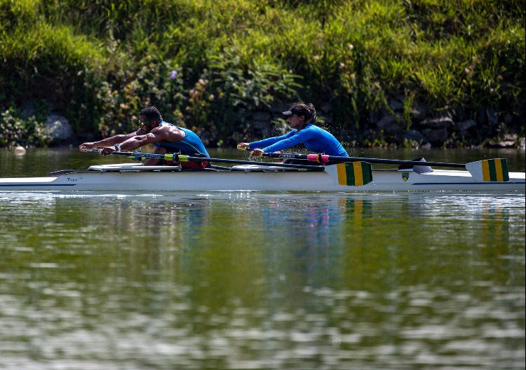 Dois atletas do remo, em embarcação dupla na cor branca, durante treino na Raia da USP. Água em primeiro plano. 