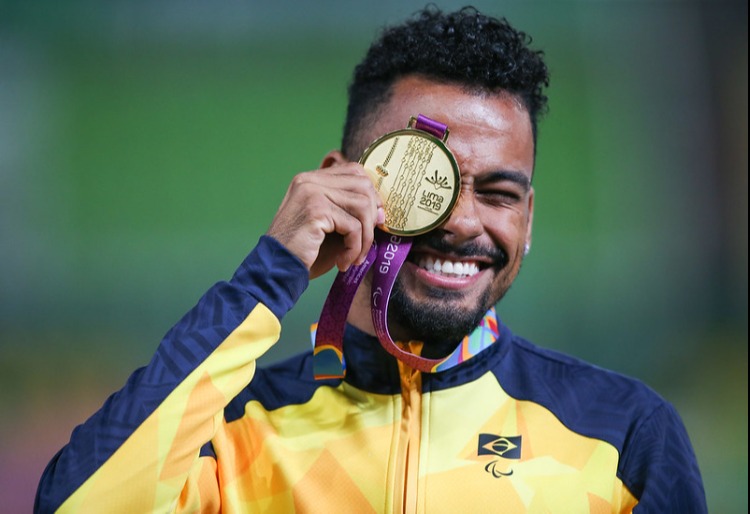 Daniel Martins, homem negro, sorrindo, segurando medalha de ouro na altura do olho e piscando o outro. Ele veste casaco do uniforme do Brasil nas cores amarela e azul escuro.