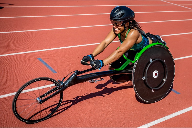 Aline Rocha, mulher negra, com capacete preto e óculos espelhado, top verde e luvas pretas na cadeira de rodas de corrida (preta com detalhes verde) na pista de atletismo