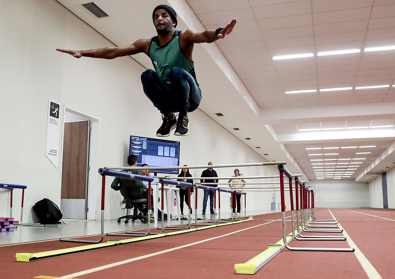 Jovem negro de uniforme verde salta obstáculo na pista interna de atletismo do CT Paralímpico, no fundo vê-se o técnico da Seleção