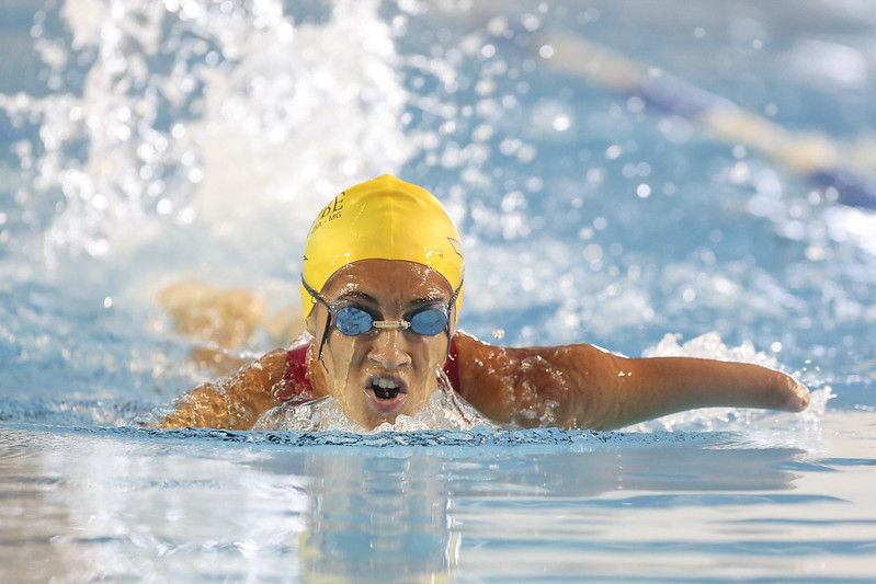 Nadadora negra e amputada dos dois braços usa touca amarela e faz respiração durante o nado borboleta na piscina do CT Paralímpico. Ela é vista de frente.